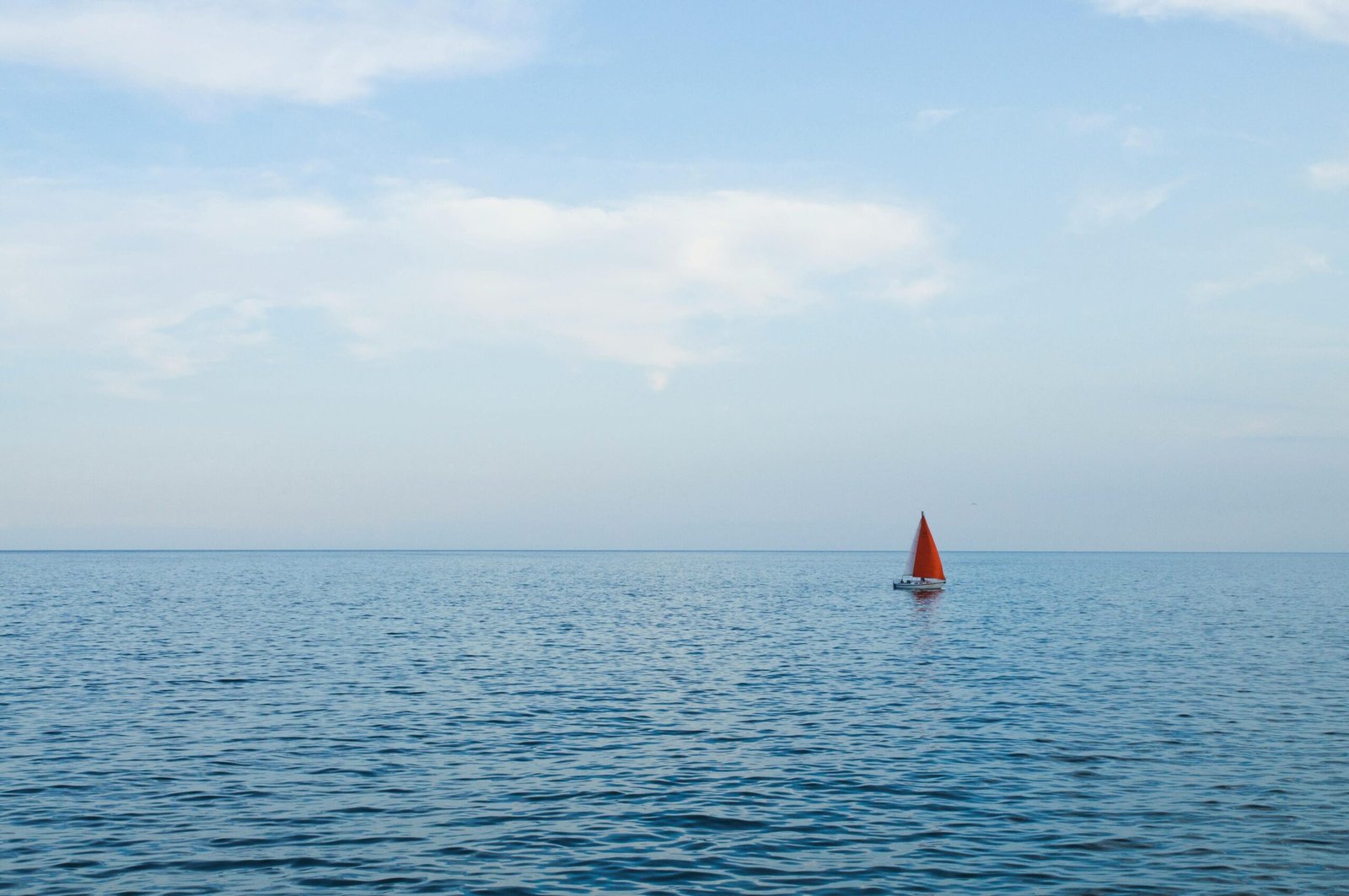 A lone sailboat with a red sail peacefully sailing on the open sea under a clear sky.